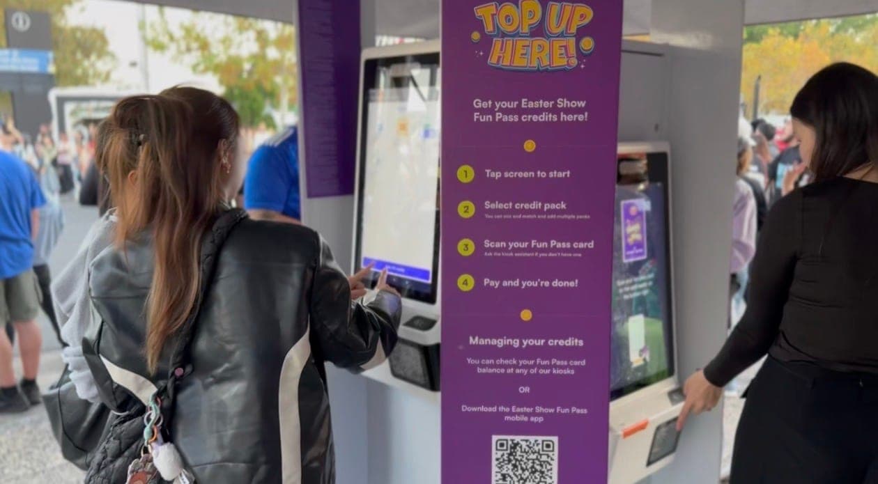 Two visitors using the Top Up Here kiosks at the Ekka with the Carnival Fun Pass branding and instructions
