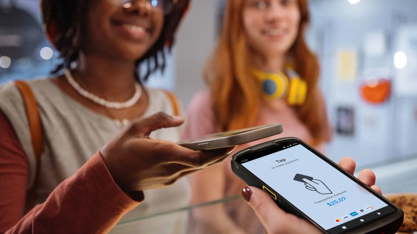 A visitor tapping their phone on a Ludo POS terminal at a venue checkout, friend smiling in the background